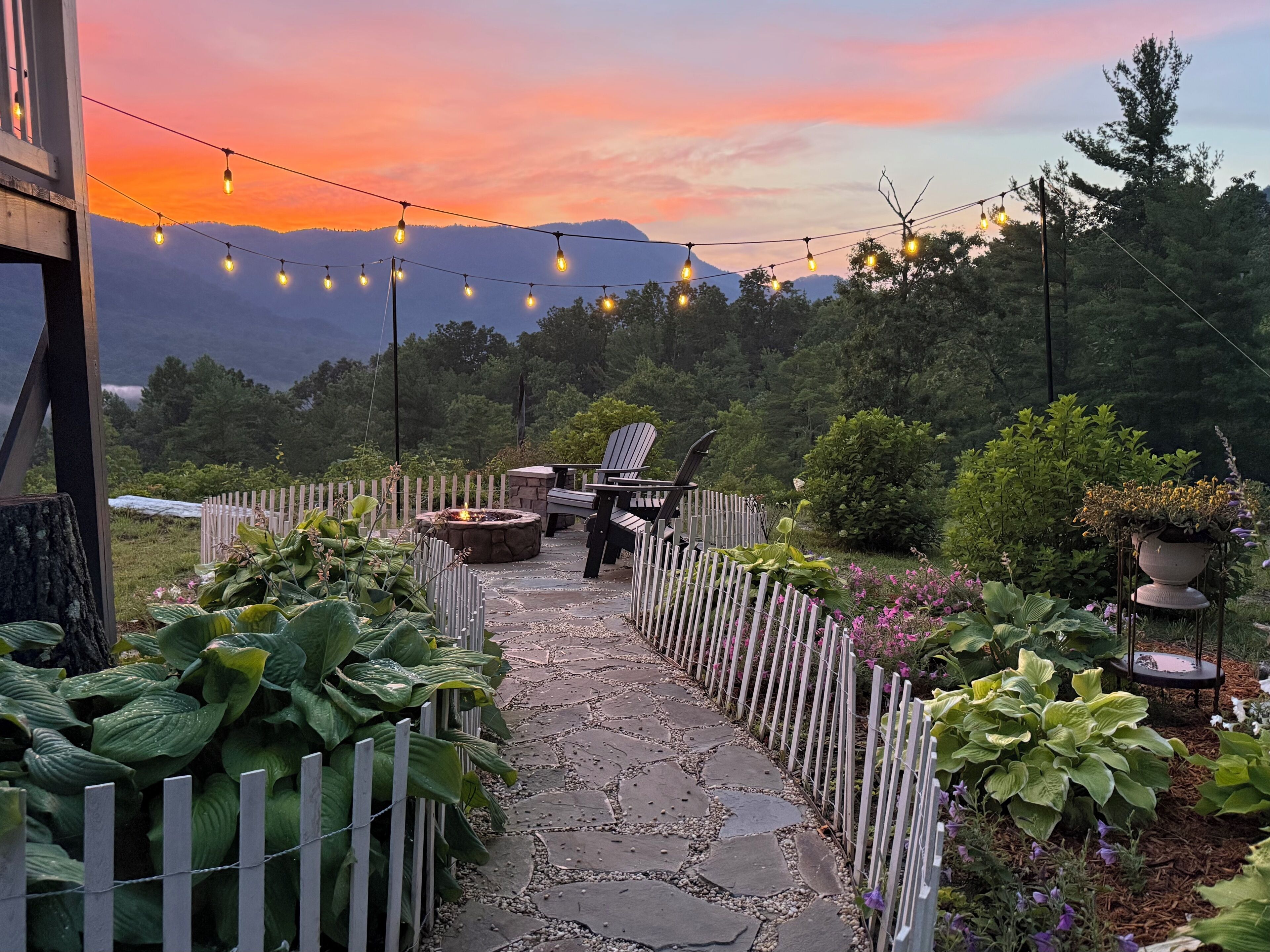 A stone pathway leading to outdoor seating with a fire pit, surrounded by lush greenery and a wooden fence.