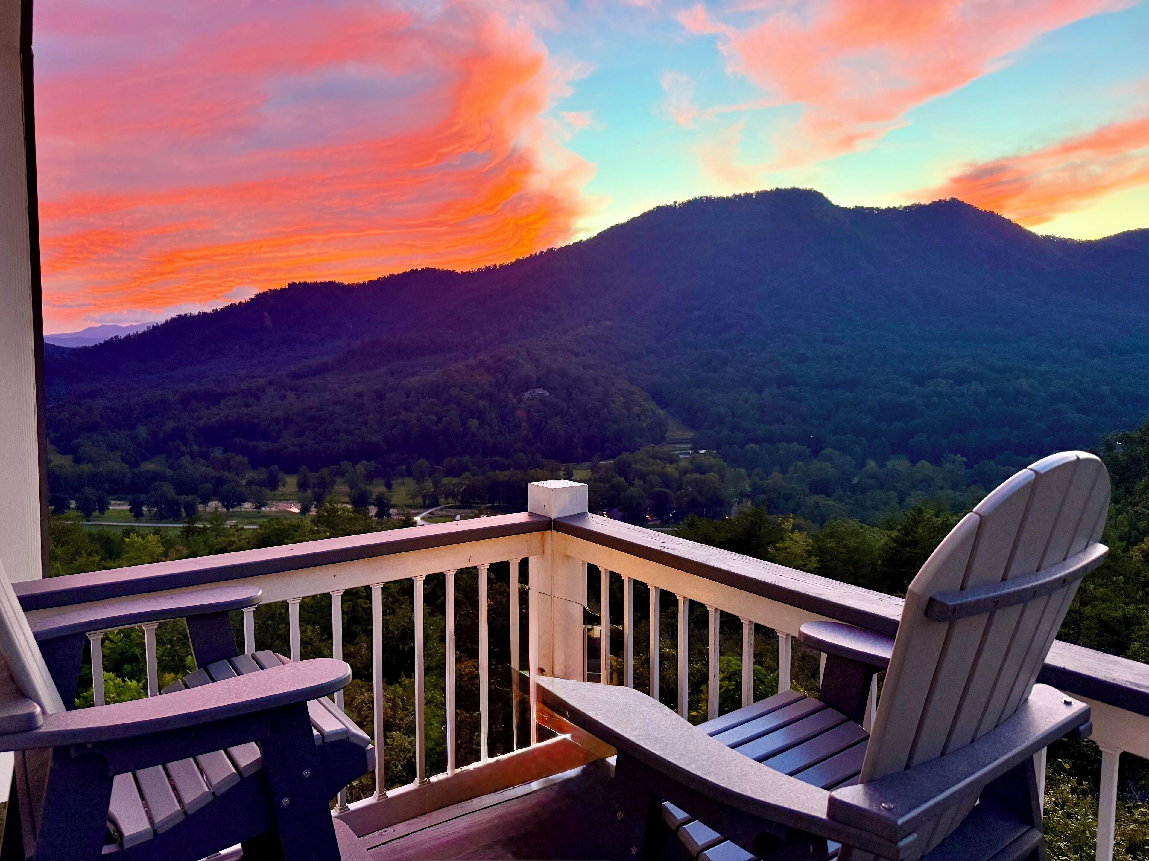Un balcón con sillas Adirondack con vista a un paisaje montañoso al atardecer.