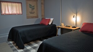 A room with two beds, a checkered rug, a framed poster, a lamp, and a small table with a hat and book. - T's Fish Camp--Crappie Are Biting—Sleeps up to Six (Warsaw)