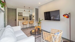 A modern living room with a white sofa, a round coffee table, and wicker chairs. A kitchen with white cabinets and a dining area with bar stools is visible in the background. - Poblado Paradise for Your Stay 2br 2bath (Medellín)