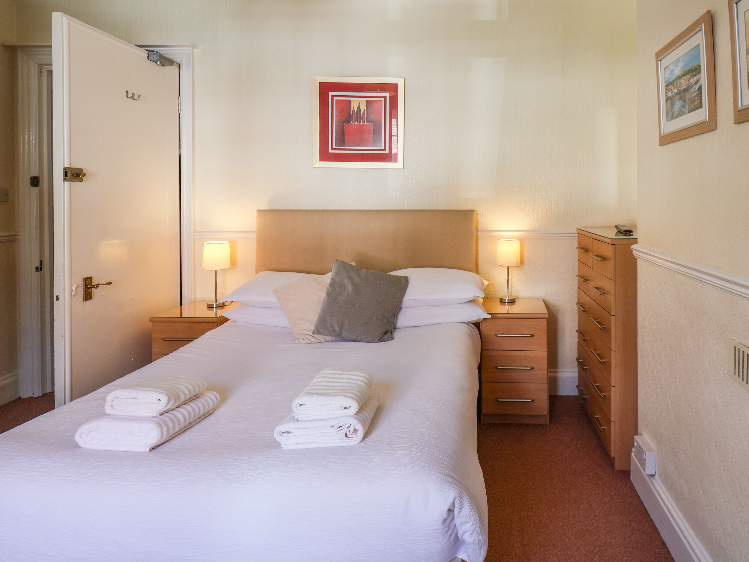 A neatly made bed with two pillows, a bedside table with a lamp, a wooden dresser, and a framed picture on the wall.