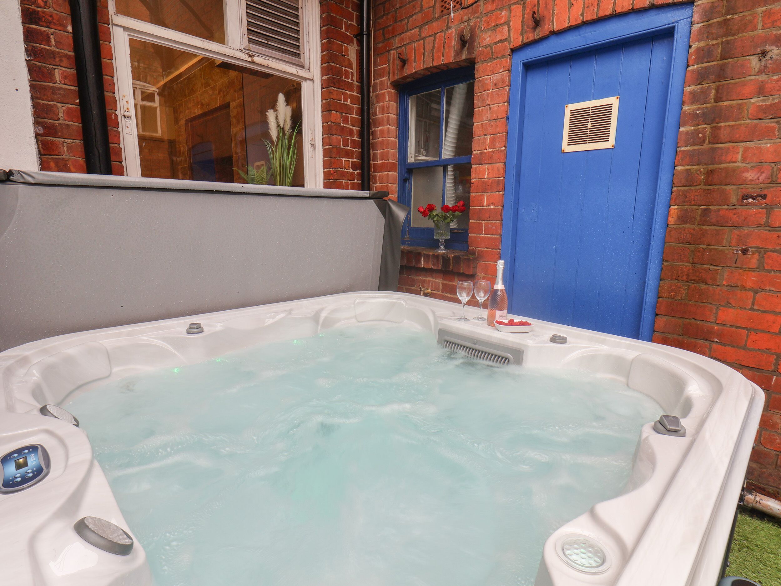 A hot tub in a backyard with a brick wall, a blue door, and a window with a view of the interior.