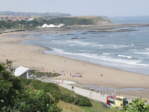 A coastal beach with a paved walkway, a lifeguard tower, and a distant view of a town. - Lonsdale Villa (Scarborough)