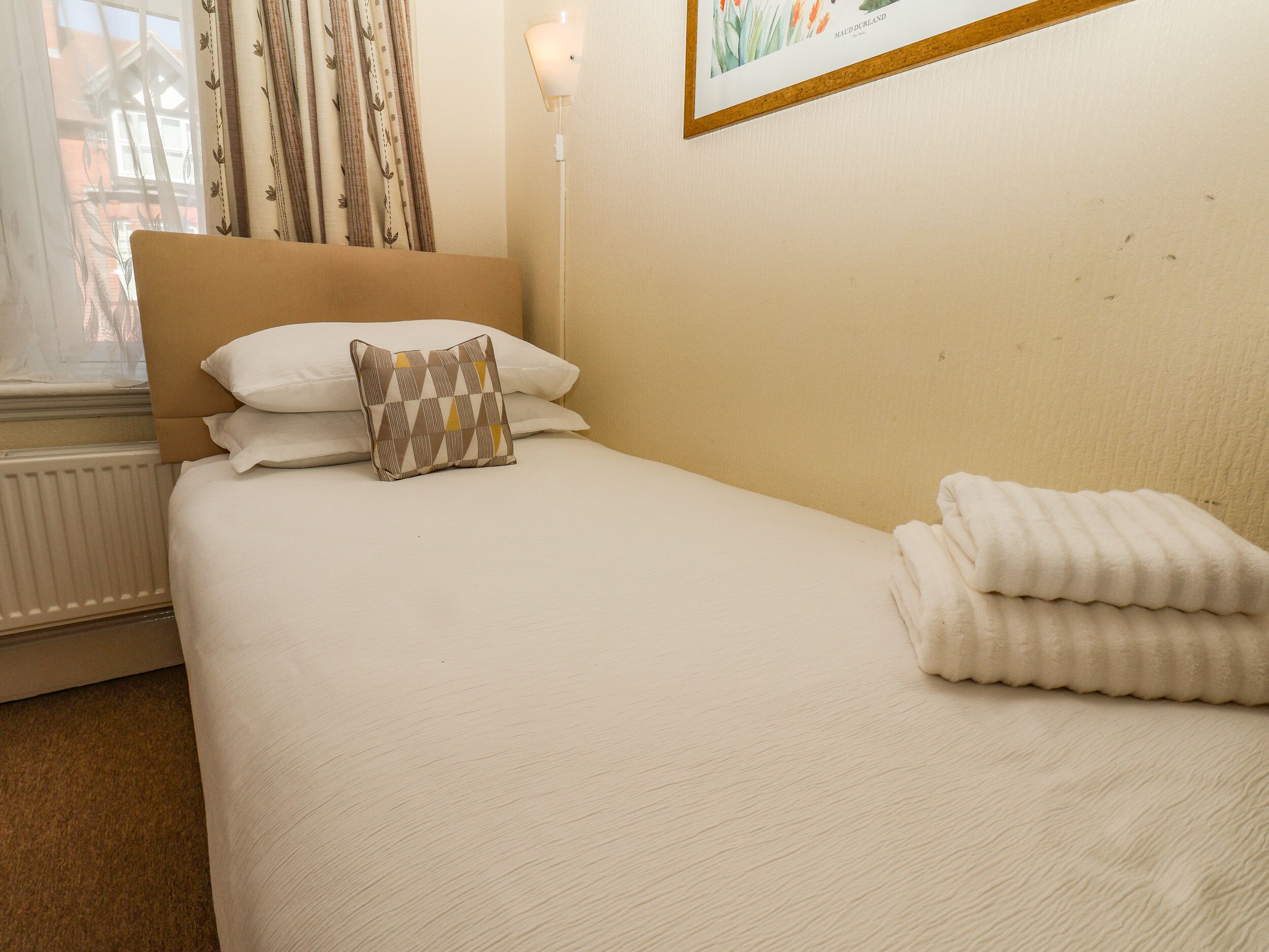 A neatly made bed with a patterned pillow and folded towels, a radiator, and a framed picture on the wall.