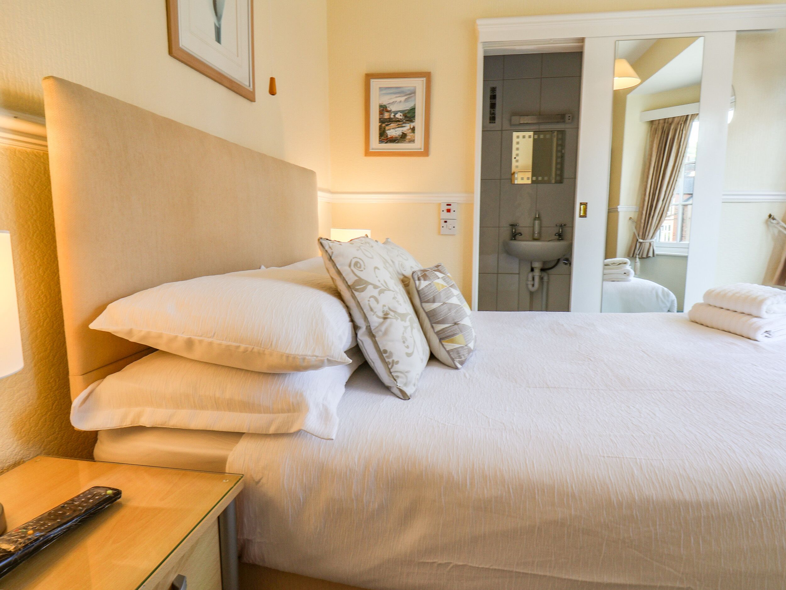 Una habitación de hotel con cama, almohadas, una mesita de noche, un control remoto, un cuadro enmarcado, y un baño con lavamanos y toallas.
