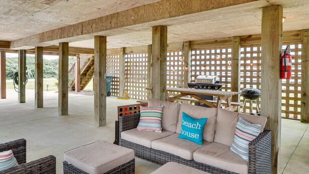 A covered patio area with a wicker sofa, cushions, and a wooden table with a grill. - Nags Head Vacation Rental w/ Hot Tub: Near Beach! (Nags Head)
