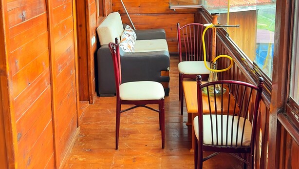 Interior de una cabaña de madera con sofá, sillas, una mesa de comedor y una ventana con vista a una casa.