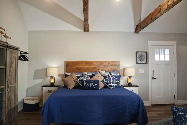 Un dormitorio con cabecera de madera, cama con ropa de cama azul, dos mesitas de noche con lámparas, una puerta y una alfombra pequeña.