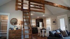A loft-style living space with a wooden beam ceiling, a dining area with white chairs, a kitchen with dark cabinets, and a staircase.