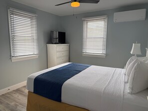 Une chambre à coucher avec un lit, deux fenêtres équipées de stores, un ventilateur de plafond, un téléviseur posé sur une commode et un climatiseur fixé au mur.