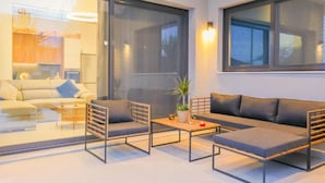 Een moderne woonkamer met een bank, twee fauteuils en een salontafel. Grote ramen en uitzicht op de keuken.