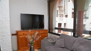 A living room with a grey sofa, a wooden cabinet, a flat-screen TV, and a window with blinds.