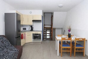 A compact kitchen with a refrigerator, microwave, and oven. There is a dining table with chairs and a vase with flowers. A sofa is visible in the foreground. - Beautiful appart with shared pool (Le Barcarès)