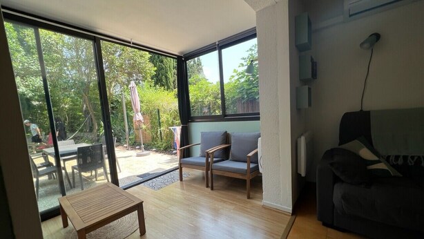 A spacious living room with large windows, a wooden table, a sofa, and a chair. - Le tinharé - studio with garden near the sea (La Ciotat)