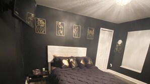 Een slaapkamer met een bed in donkere tinten, een wit hoofdbord, een houten vloer en een plafondlamp.