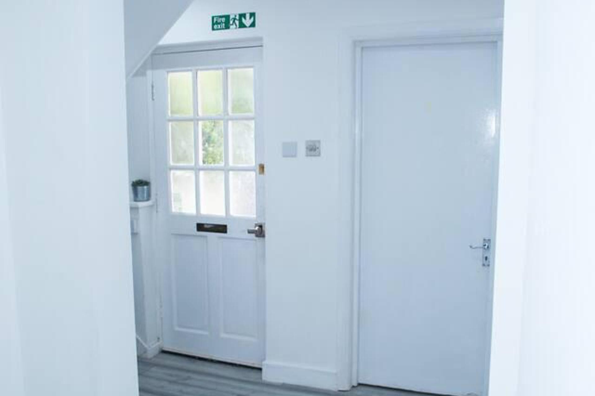 A white door with a window, a fire exit sign above, and a small shelf with a container.