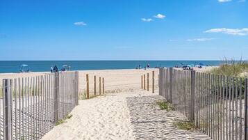 Ein sandiger Strandweg führt zum Meer, begleitet von einem Holzzaun auf der einen Seite und Strandsonnenschirmen auf der anderen.