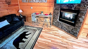 A cozy cabin interior with a wooden wall, a mounted TV, a fireplace, a sofa, a small table with stools, and a rug with a bear silhouette. - Snuggle Bear: Cozy, Enchanting Mtn. Retreat Haven. New Renovated & deep cleaned! (Gatlinburg)