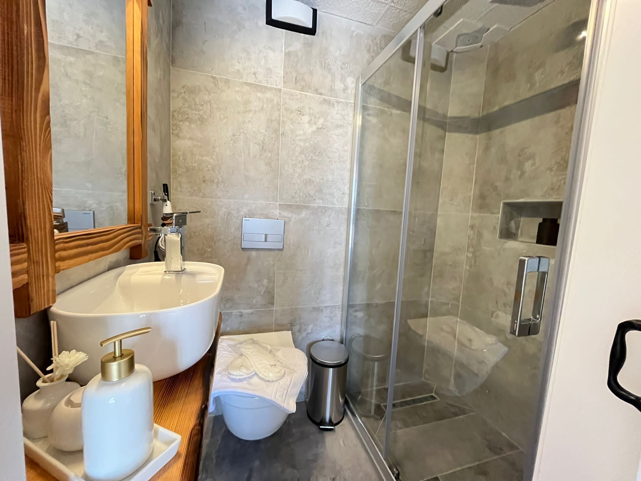 Une salle de bain avec un grand lavabo blanc, un miroir, une cabine de douche en verre et une petite poubelle.