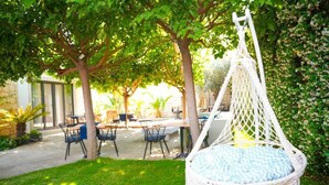 A hammock chair with a patterned cushion in a garden area with tables and chairs, surrounded by greenery and a building in the background. - Armi Alaçatı (Cesme)