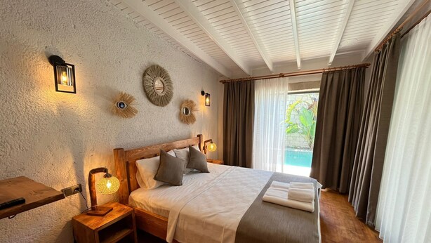 A bedroom with a wooden bed, bedside lamps, a small wooden nightstand, and a view of a swimming pool through the window. - Armi Alaçatı (Cesme)