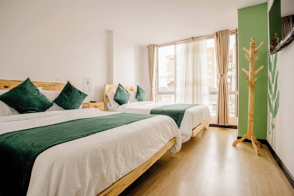A hotel room with two beds, wooden headboards, green bedding, a wooden floor, and a view of buildings through the window.