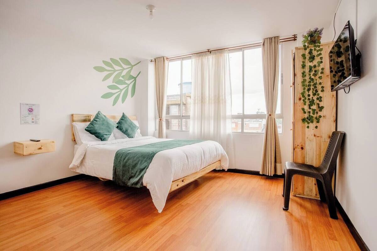 A bedroom with a wooden bed, a chair, a TV, and a window with curtains.