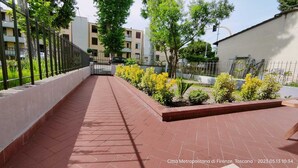 Un sendero pavimentado con diseño de ladrillo, delimitado por un muro bajo y una cerca de metal. Hay plantas y arbustos pequeños a lo largo del muro.