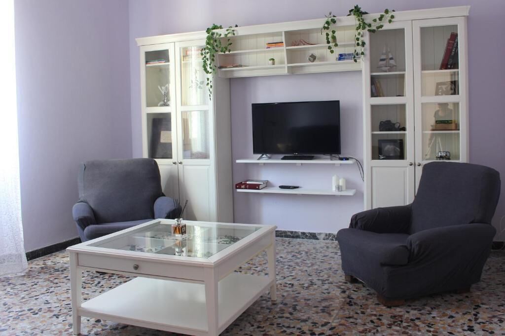 Un soggiorno con due poltrone, un tavolino in vetro, una TV a schermo piatto e un mobile bianco con ripiani.