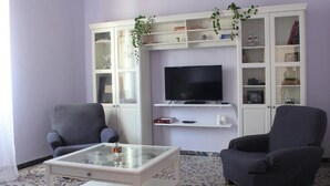 Una sala de estar con dos sillones, una mesa de centro de vidrio, un televisor de pantalla plana y un mueble blanco con estantes.