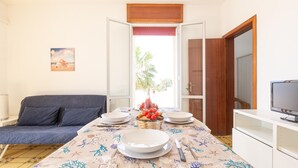 Ein Zimmer mit einem Bett, einem Esstisch gedeckt mit Tellern, einer Obstkorb, einem Fernseher auf einem Schrank und einem Fenster mit Blick auf Palmen.