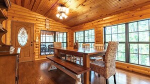 Interior de una cabaña de madera con comedor, ventanas amplias y un piano.