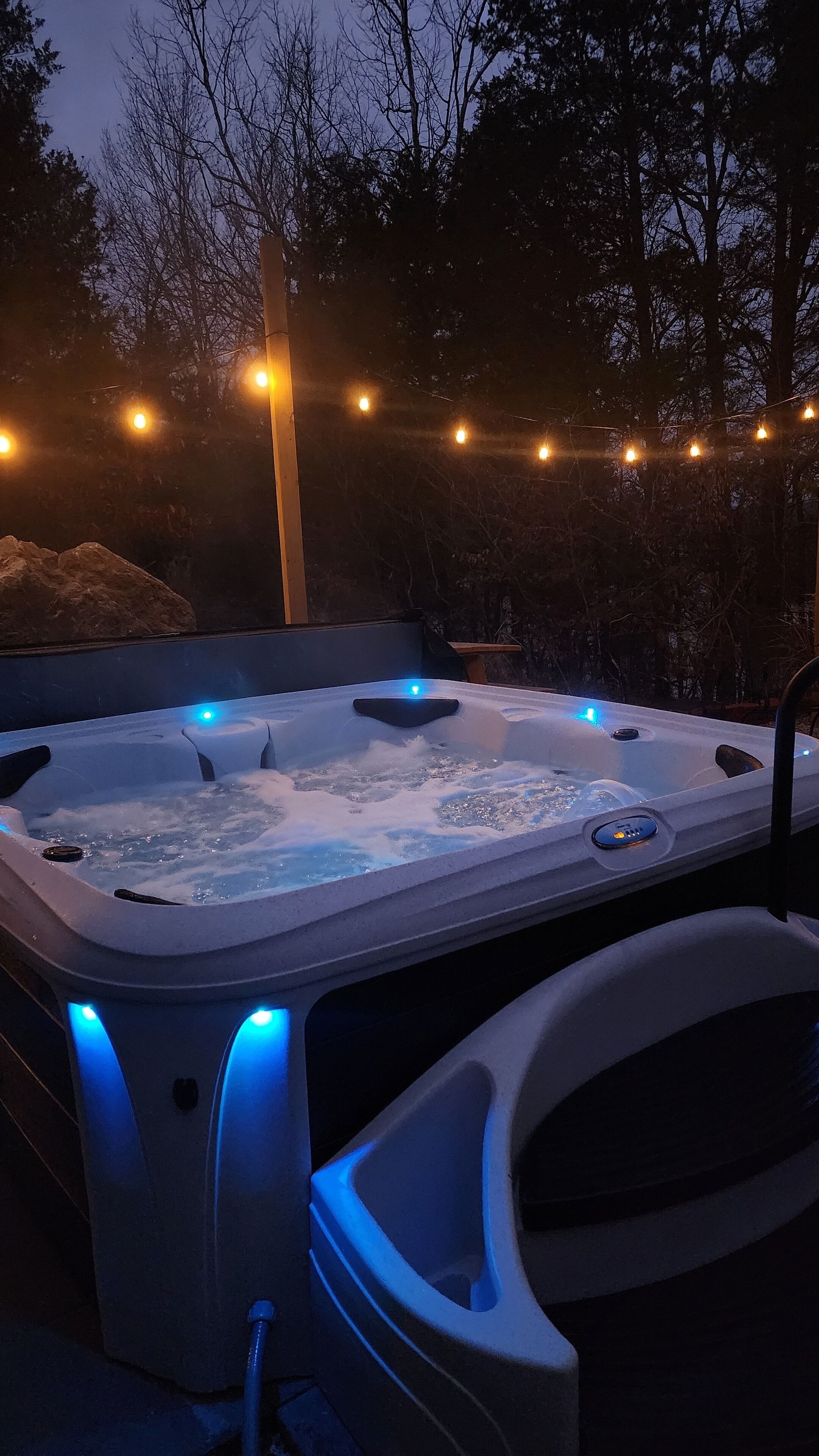 En utomhusbubbelpool med blått LED-ljus, omgiven av träd och ljusslingor.