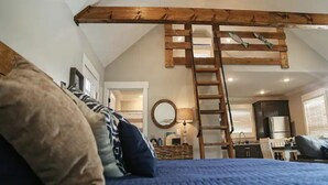 Un dormitorio con un entrepiso para dormir, una escalera de madera y una cocina al fondo.