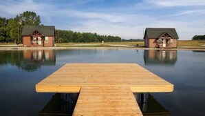 Un muelle de madera que se adentra en un lago tranquilo, con dos cabañas en la otra orilla.