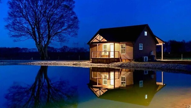 Una cabaña de madera junto a un lago tranquilo por la noche, con su reflejo visible.