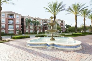 Stroll through the serene landscaped area, surrounded by a central fountain and palm trees, with a multi-story building in the background.
