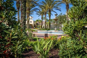 Surrounded by lush greenery and palm trees, this serene oasis features a central fountain and a classic building in the background.
