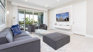Een moderne woonkamer met een grijze bank, een poef en fauteuils, een glazen salontafel en een grote flatscreen televisie aan de muur.