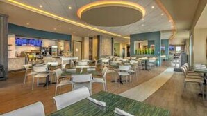 Een ruime hotel eetzaal met meerdere tafels en stoelen, een centrale bar en een modern plafondontwerp.