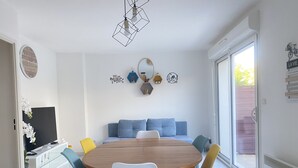 A dining area with a round wooden table surrounded by colorful chairs, a blue sofa, and decorative wall art. - Maison Sécurisée à 2km de la Plage et du Centre-ville (Soulac-sur-Mer)