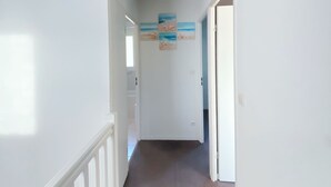 A hallway with white walls, a brown floor, and a ceiling light. There are three paintings on the wall depicting beach scenes. - Maison Sécurisée à 2km de la Plage et du Centre-ville (Soulac-sur-Mer)