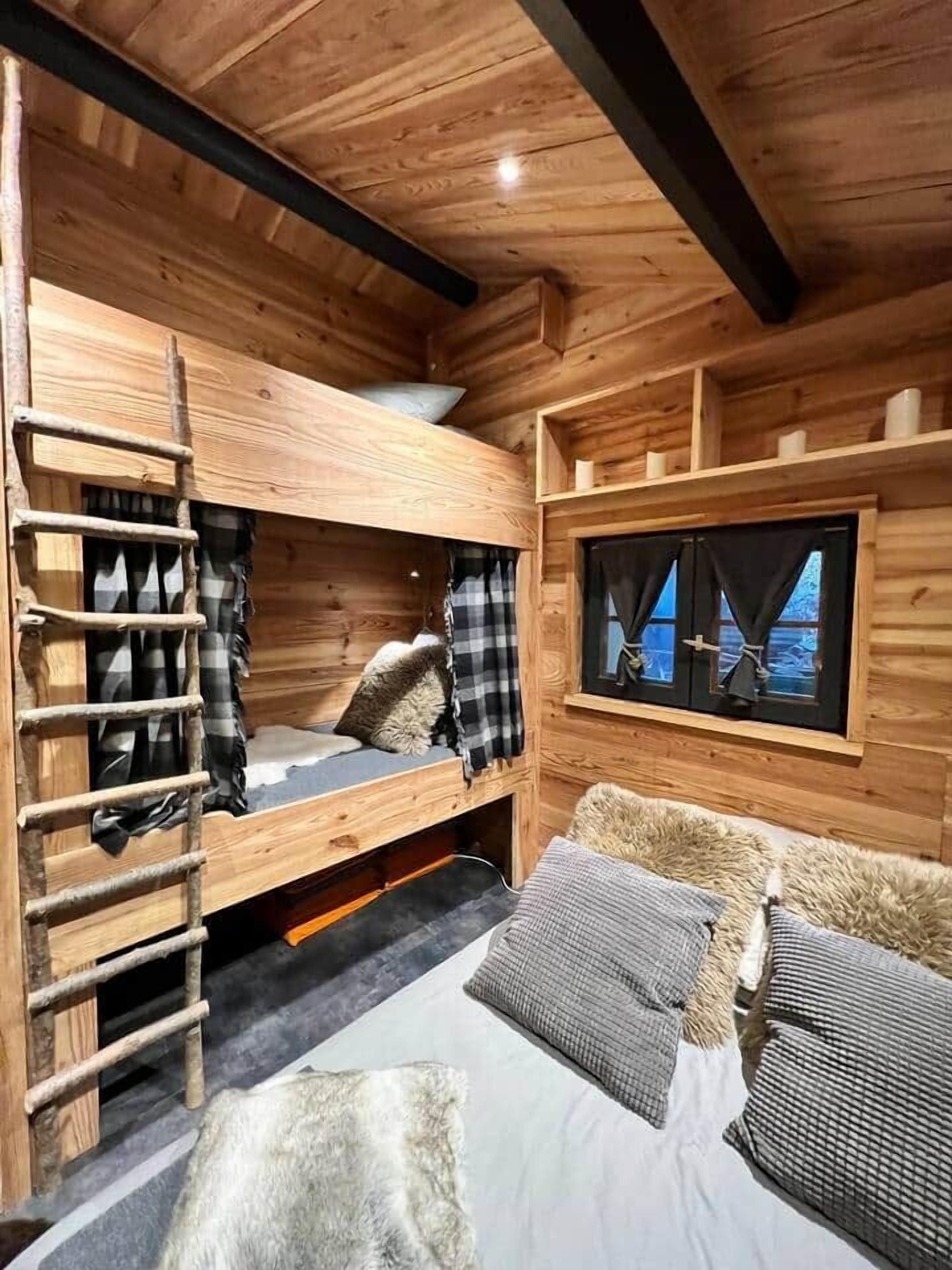 Une cabane en bois avec des lits superposés, une échelle, une fenêtre avec des rideaux et un canapé avec des coussins.