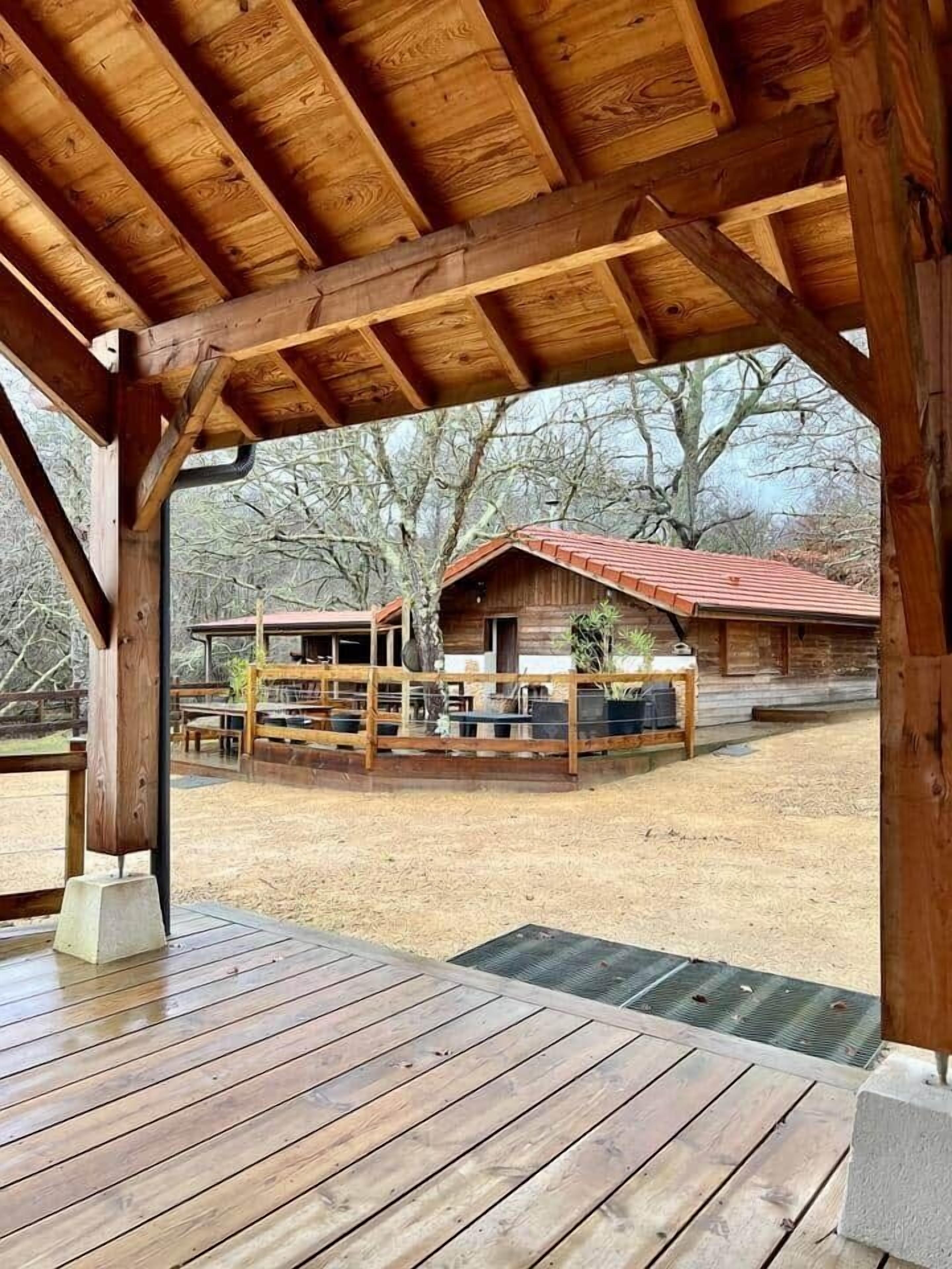 Une terrasse en bois dotée d’un espace couvert, un bâtiment rustique avec un toit rouge et une zone extérieure clôturée.