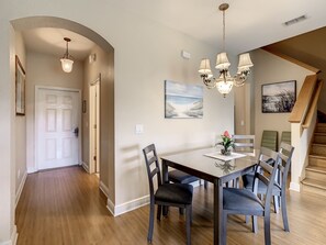 A dining area with a wooden table, six chairs, and a chandelier. There is a painting on the wall and a staircase in the background.