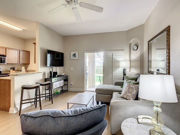A modern living room with a grey sofa, a coffee table, and a TV. There is a kitchen area with cabinets and a microwave.