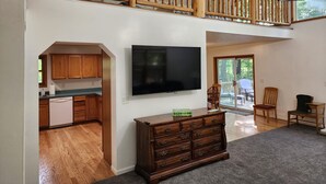 A spacious living area with a wooden staircase, a flat-screen TV, a wooden dresser, and a view of the outdoors through a large window.