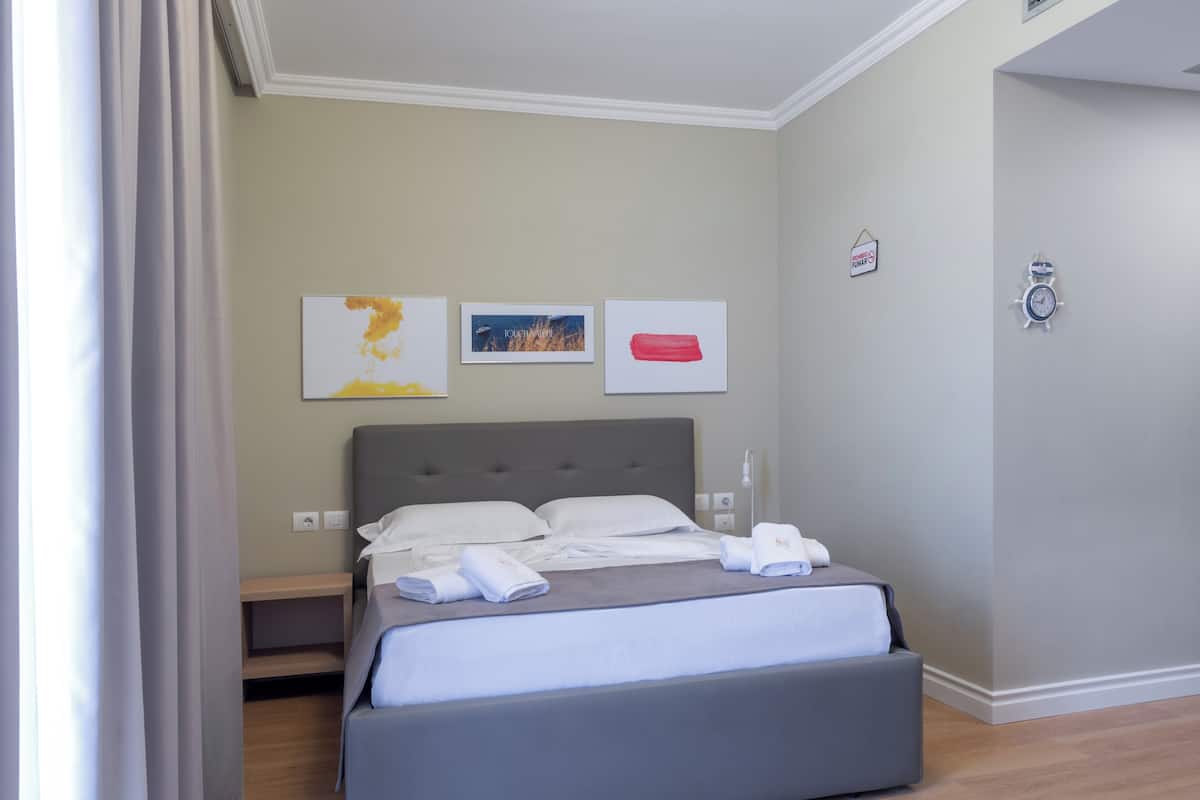 Ein ordentlich bezogenes Bett mit weißen Leinen, ein grauer Kopfteil und ein Nachttisch mit einer Lampe. Darüber an der Wand hängen drei gerahmte Kunstwerke.