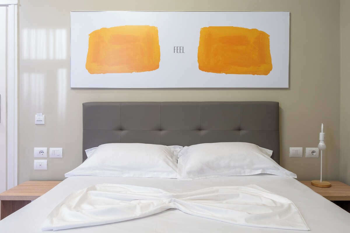 Una cama bien hecha con sábanas blancas, un cabecero tapizado en gris y dos grandes cuadros abstractos en la pared.