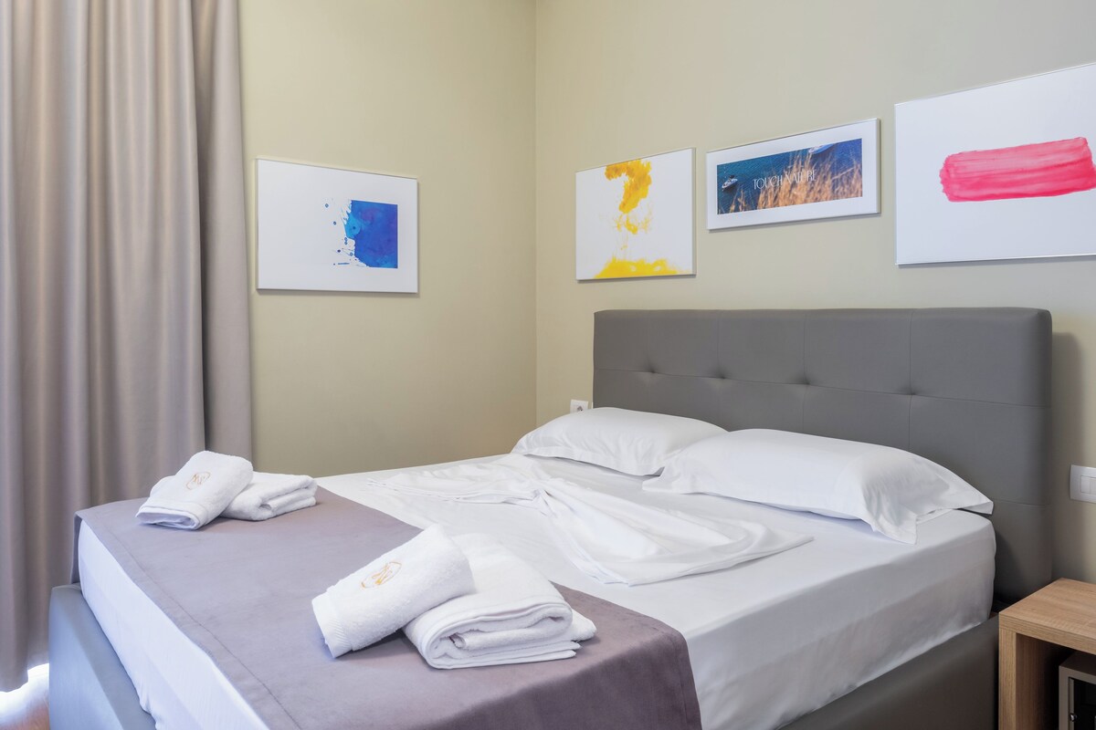Una cama bien hecha con sábanas y toallas blancas, un cabecero gris y una mesita de noche de madera.